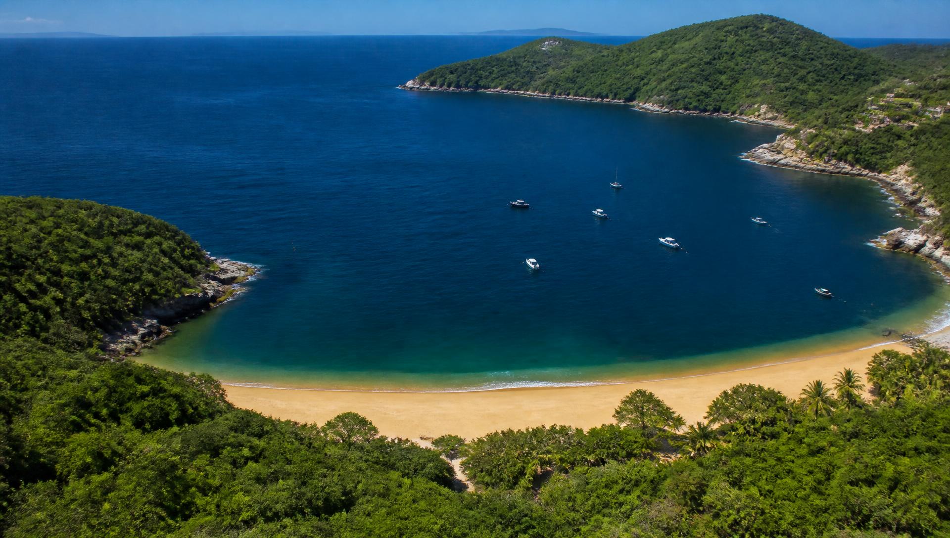 Vista aérea de una bahía de Huatulco, Oaxaca, con mar azul profundo y playa de arena dorada